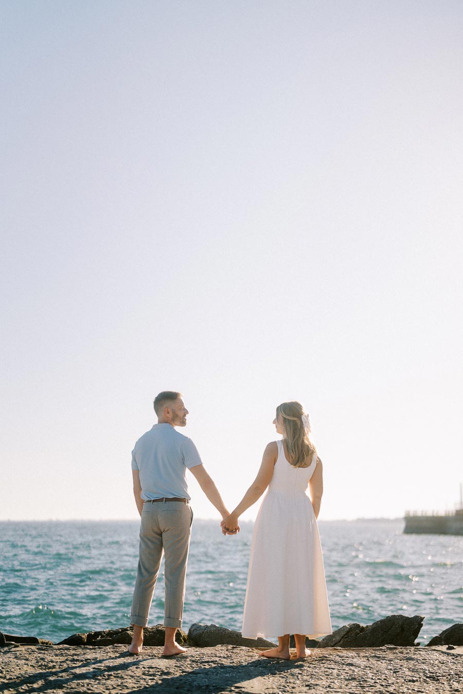 A couple holding hands stands barefoot on a rocky shore, looking out at the sea under a clear blue sky.