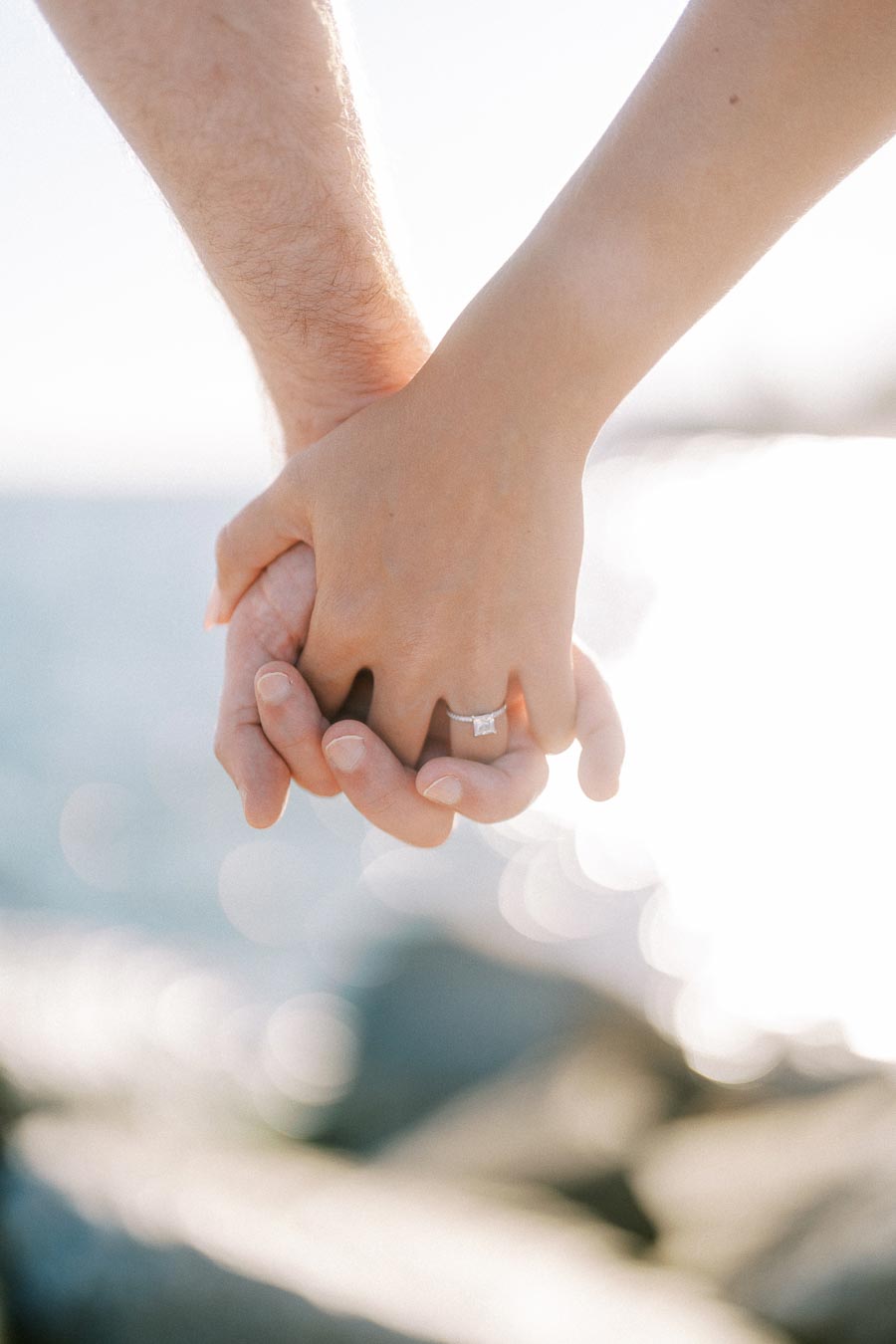 Couple holding hands near the ocean, with a focus on an engagement ring sparkling in the sunlight, symbolizing love and commitment.