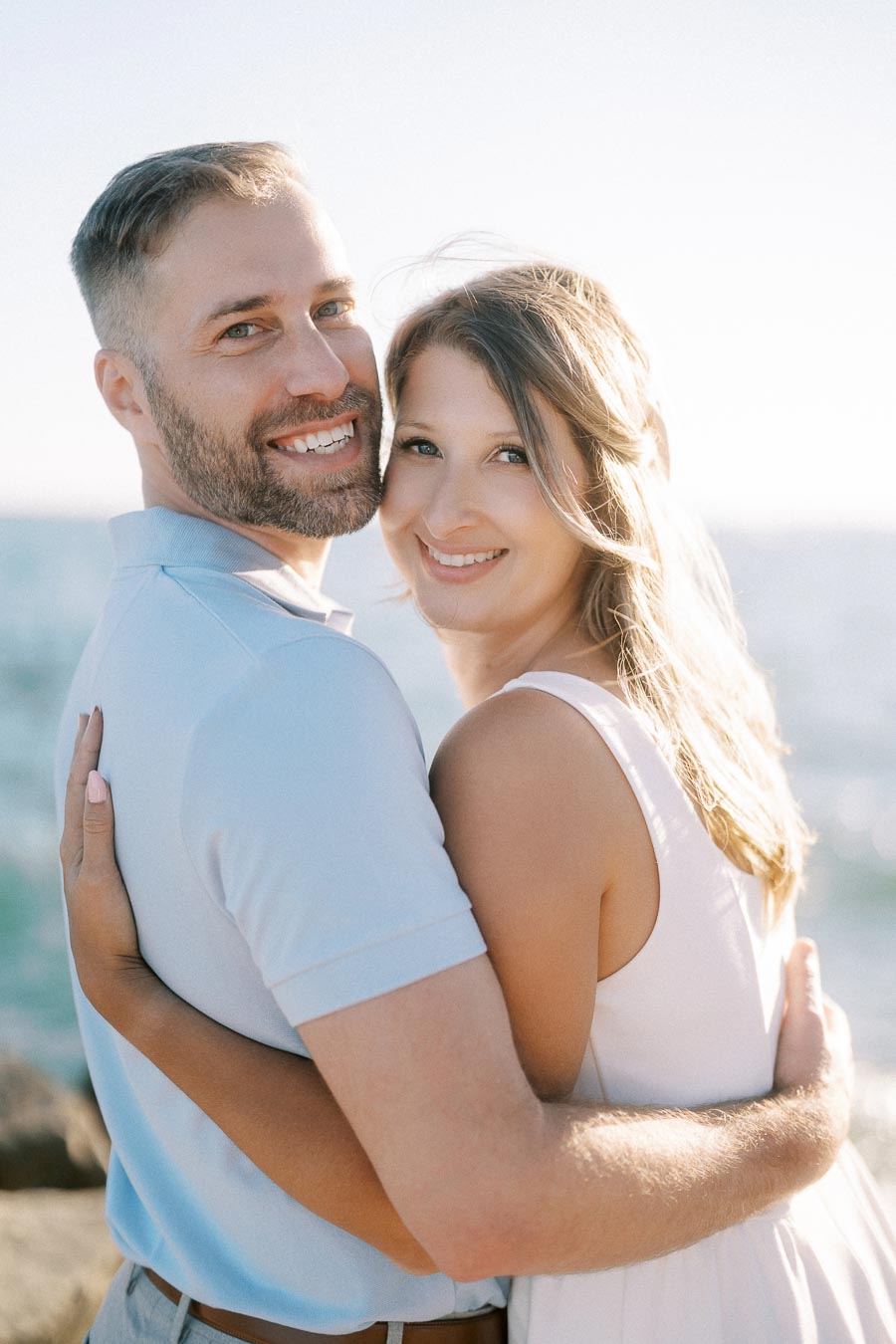 A happy couple embracing by the ocean on a sunny day, with the man in a light blue shirt and the woman in a white dress, both smiling at the camera.