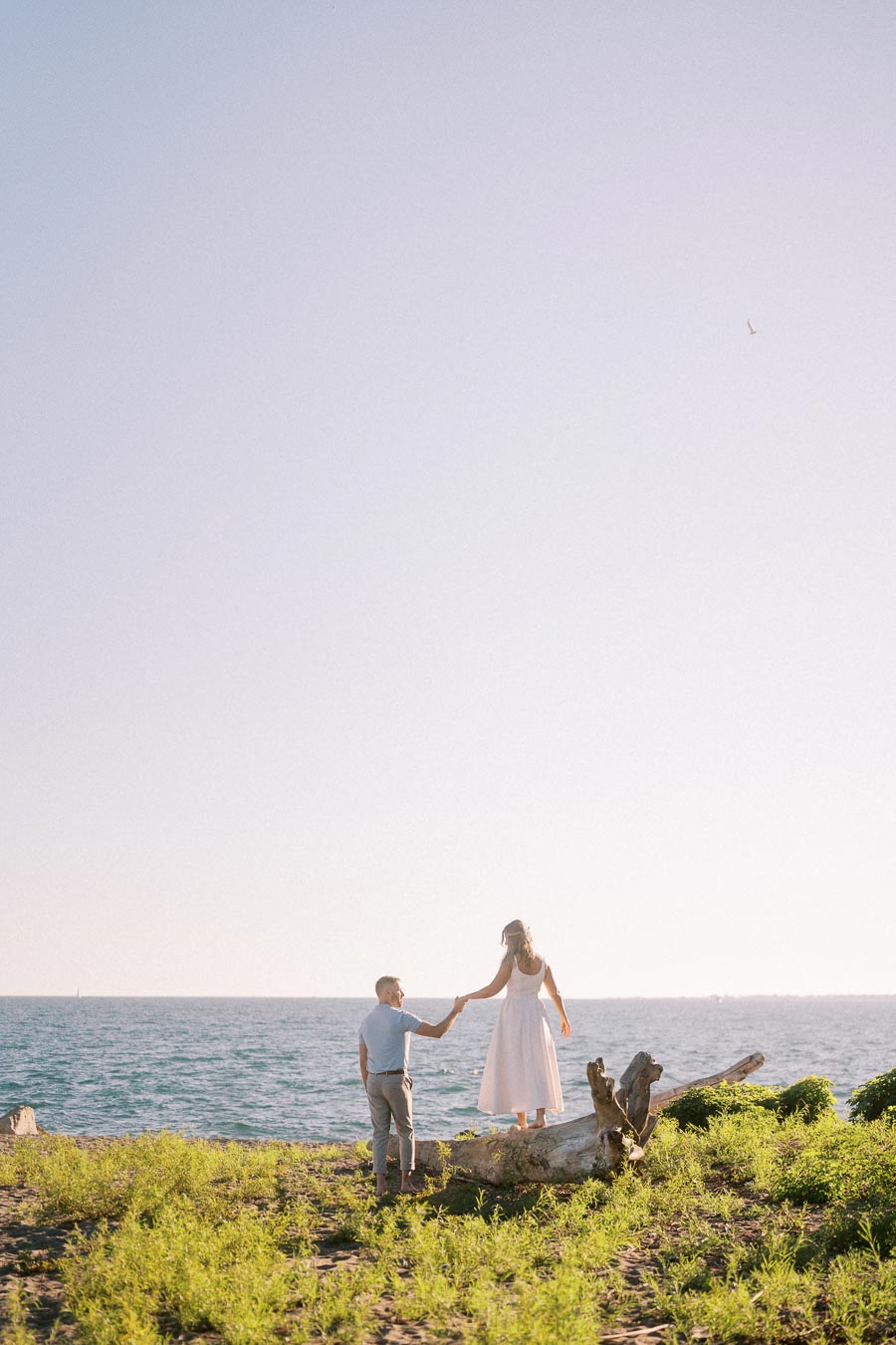 A couple enjoys a serene moment by the ocean, with the man helping the woman balance on a driftwood log against a clear sky, embodying romance and adventure in nature.