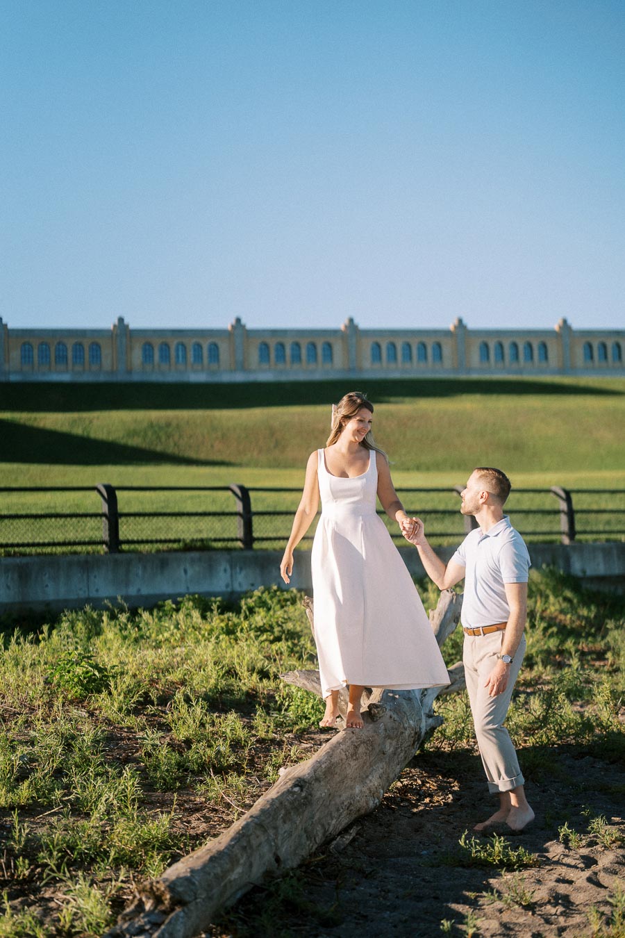 A couple enjoys a sunny day outdoors, with a woman in a white dress balancing on a log, assisted by a man holding her hand, in front of a historic building and clear blue sky.