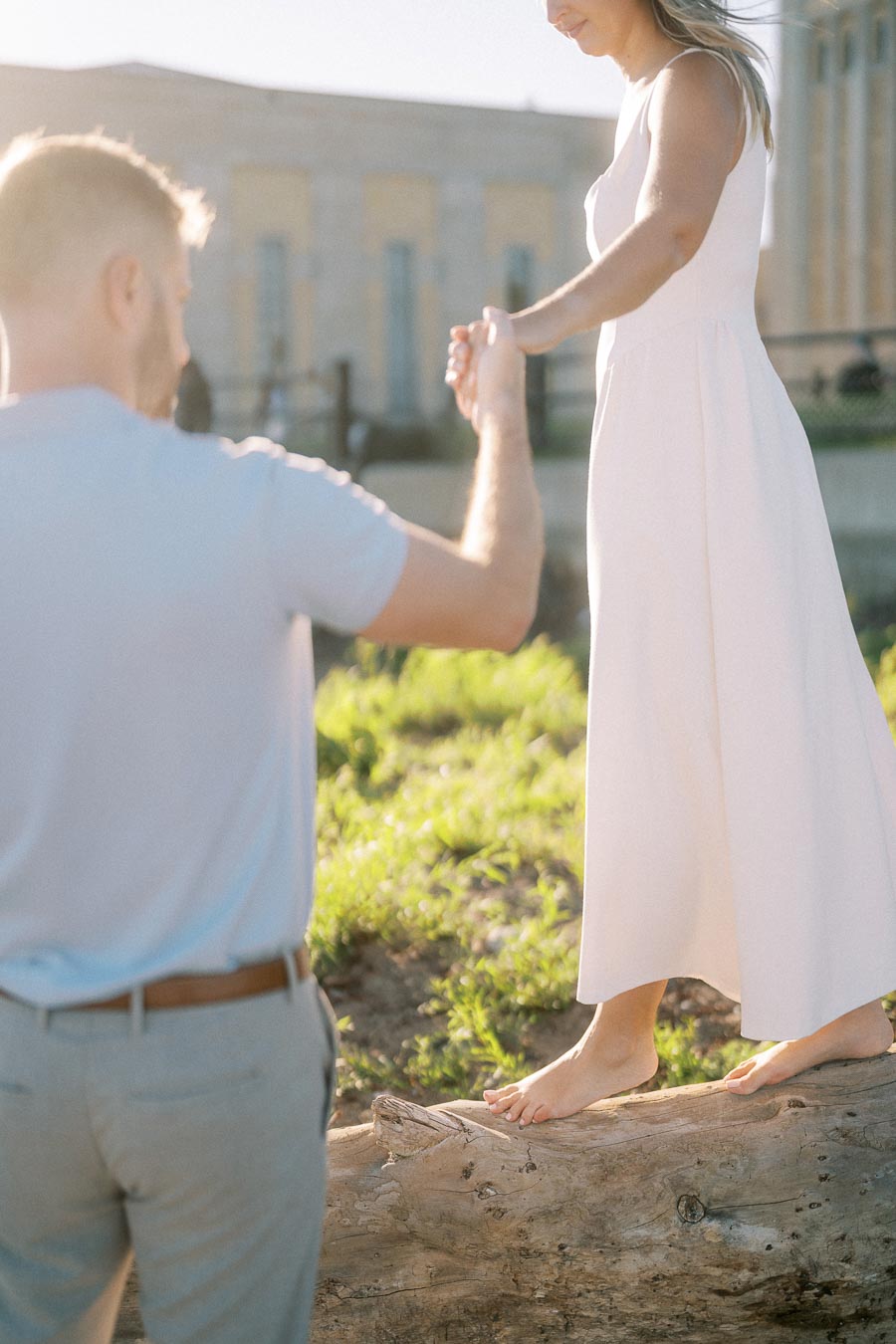 A woman in a white dress balances barefoot on a tree log in a sunny outdoor setting, while a man in a light shirt offers support by holding her hand.
