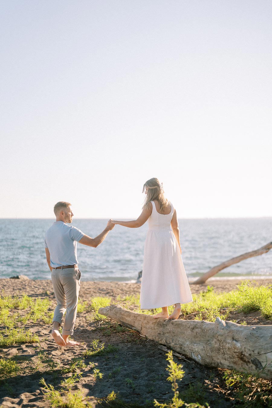 A couple walking on a sandy beach with a scenic ocean view in the background, surrounded by greenery and driftwood, under a sunny blue sky.