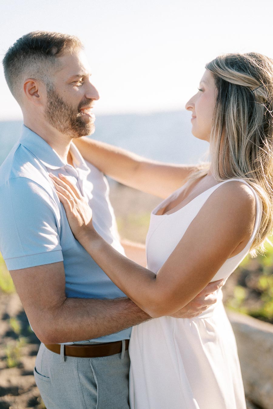 Couple embracing outdoors with a scenic ocean view, bathed in sunlight, showcasing a romantic moment.