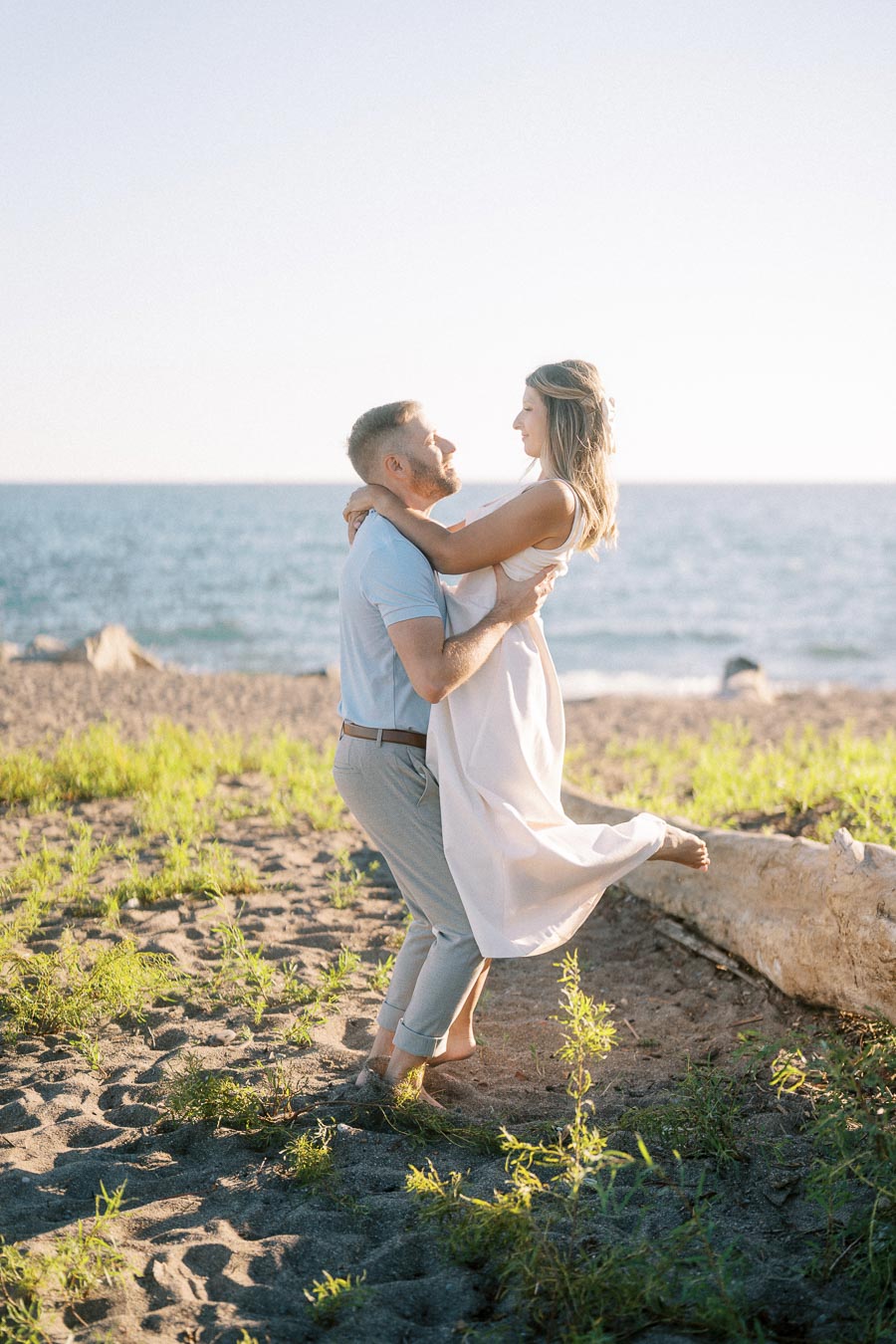 A couple embracing on a sunny beach, with the sea in the background. The woman wears a flowing white dress, and the man is in casual attire. Romantic beach setting with warm sunlight.
