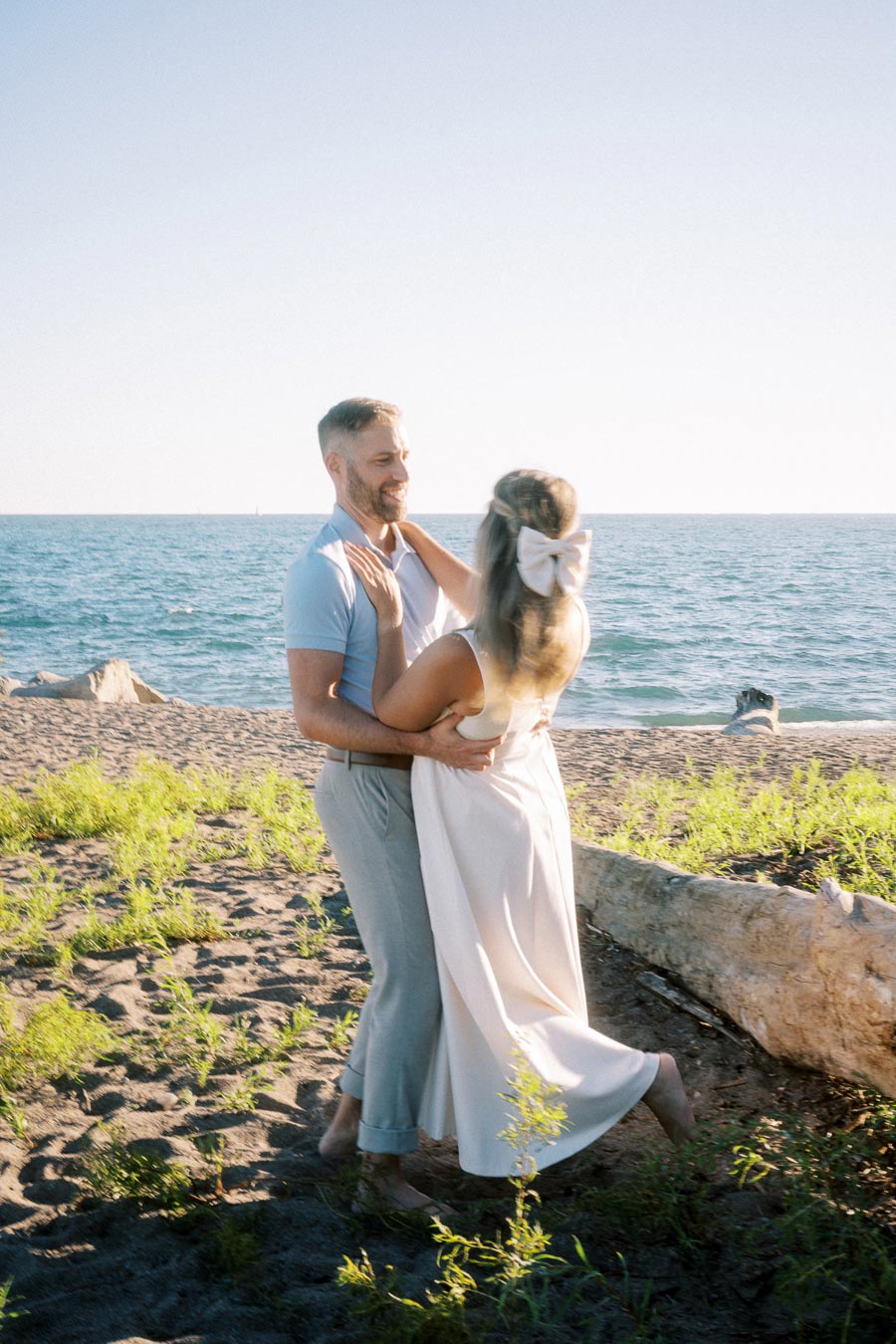 A couple embracing on a sunny beach with clear blue skies in the background, surrounded by sand and greenery near the ocean shoreline.