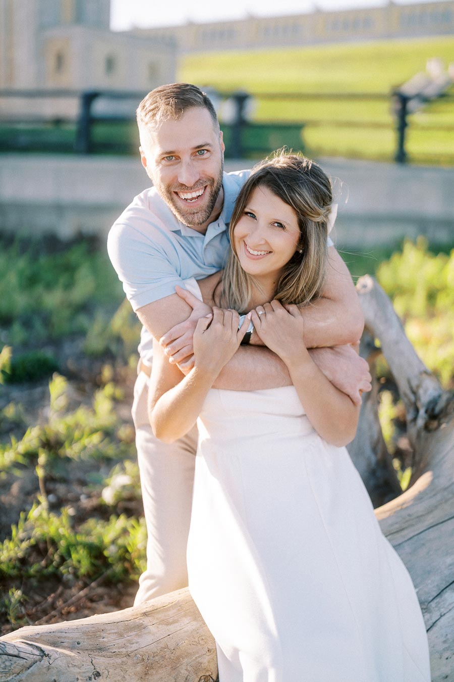 A happy couple embracing and smiling while standing outdoors in a sunlit park setting, with greenery in the background and a wooden log in the foreground.