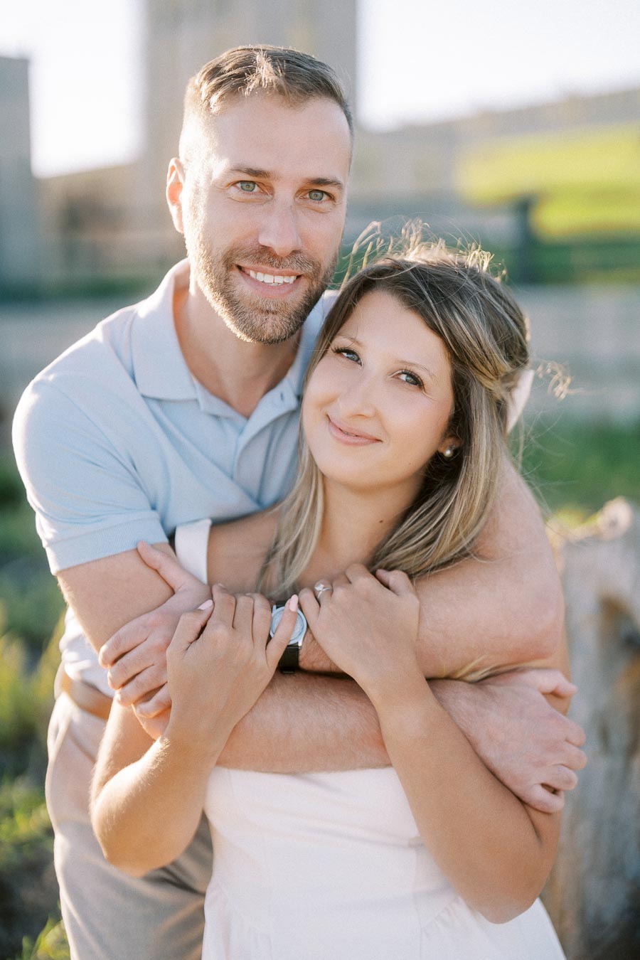 Happy couple embracing outdoors with green background, both smiling and enjoying a sunny day.