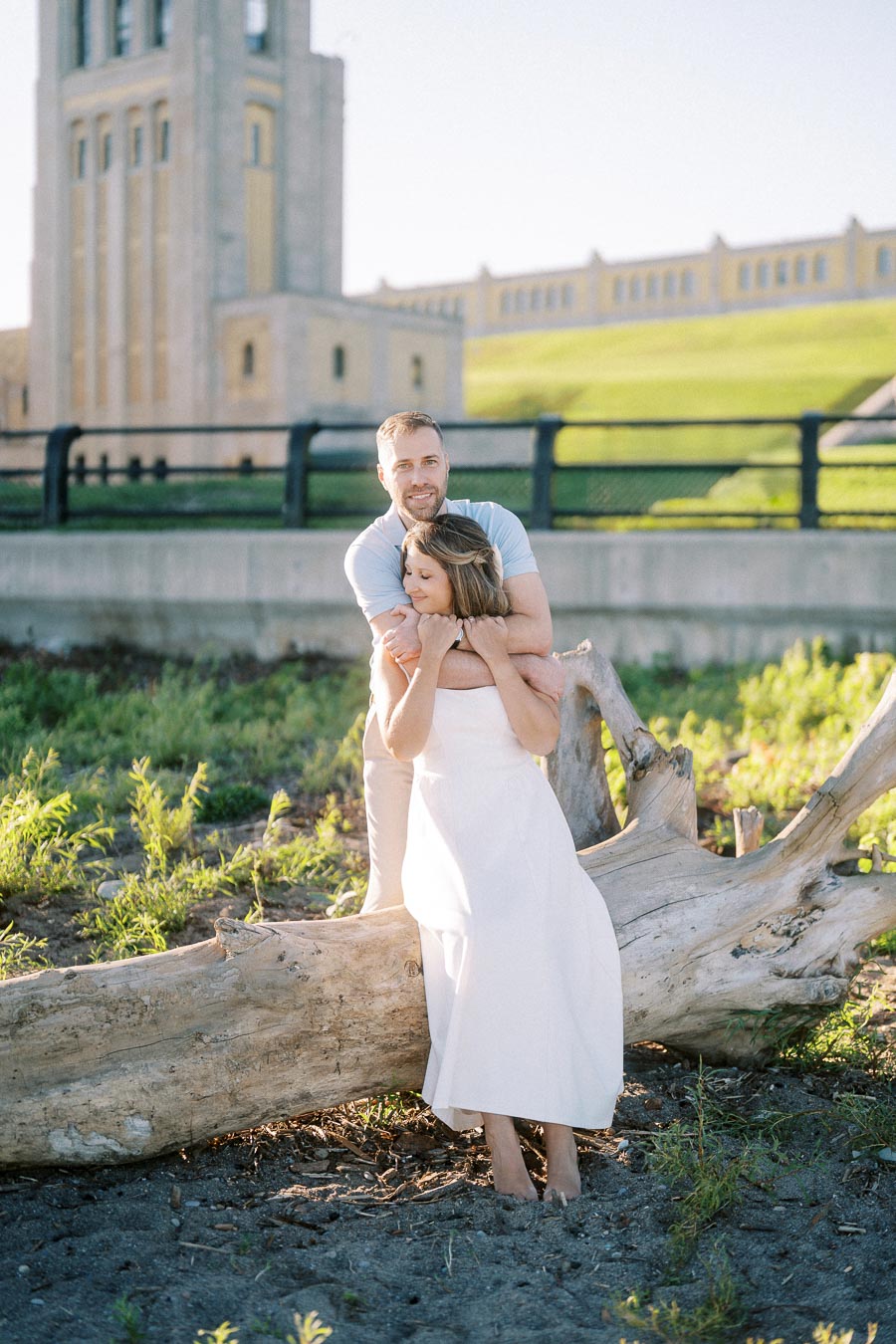A couple embraces lovingly in a sunlit park setting, with a tall, historic building in the background and greenery surrounding them.