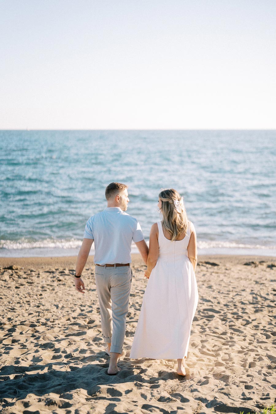A couple holding hands while walking barefoot on a sandy beach, with the ocean and clear sky in the background.