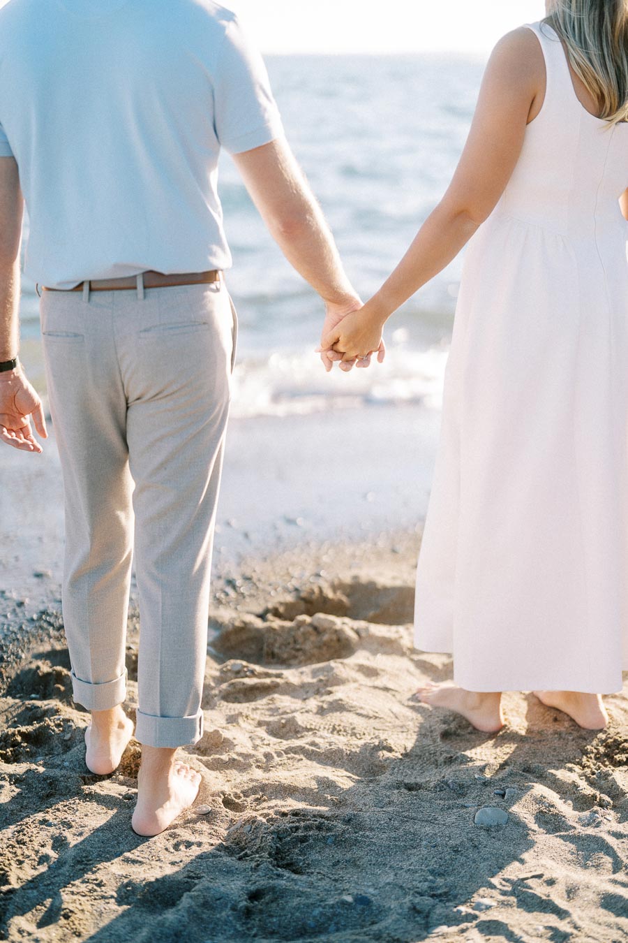 Couple holding hands on a sandy beach, wearing summer clothes and facing the ocean.