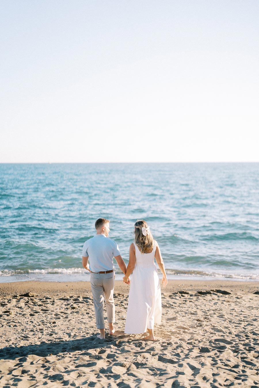 A couple holding hands, walking barefoot on a sandy beach towards the ocean, under a clear blue sky.