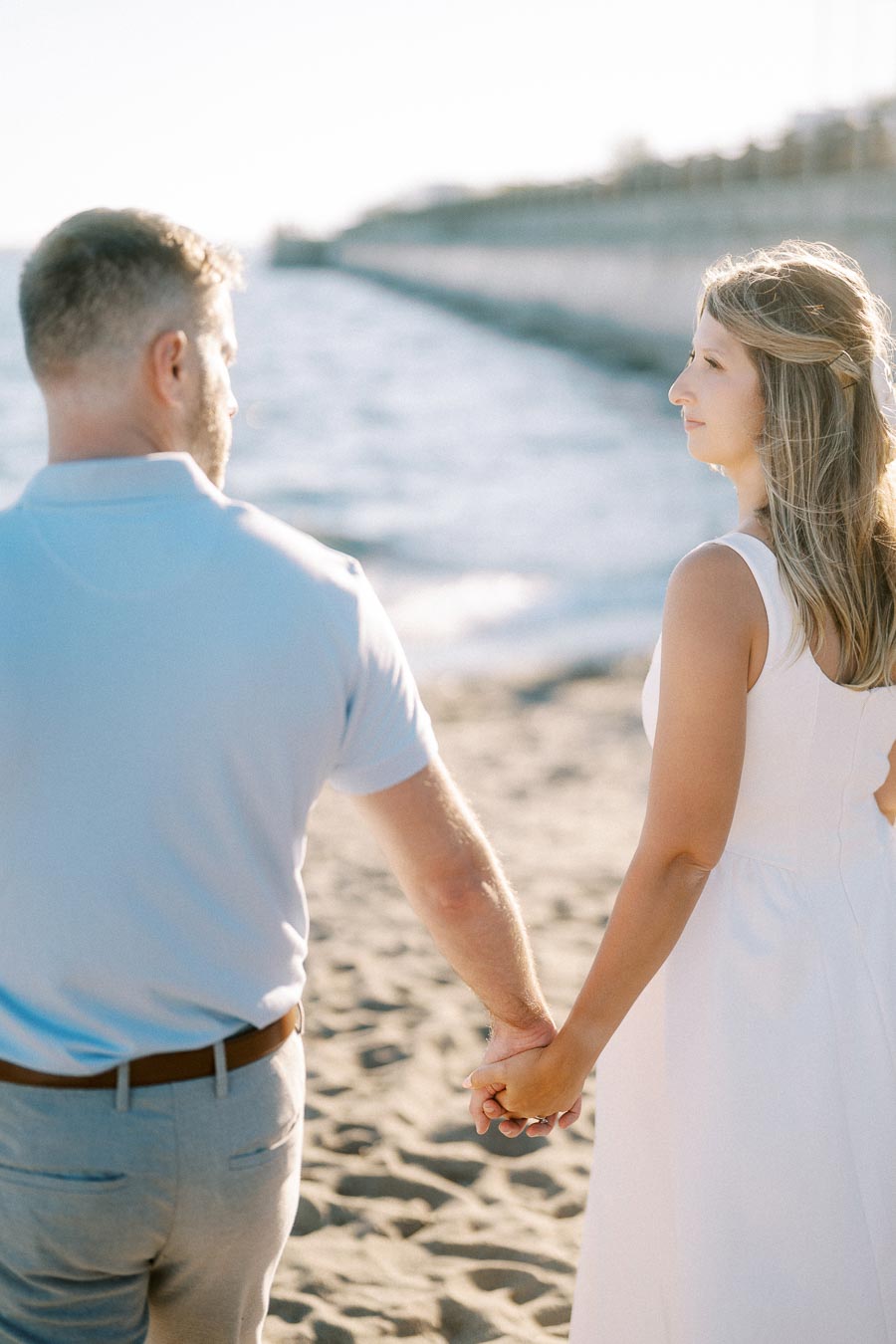 A couple holding hands on a sunny beach, with a calm ocean in the background and a pier extending into the distance.