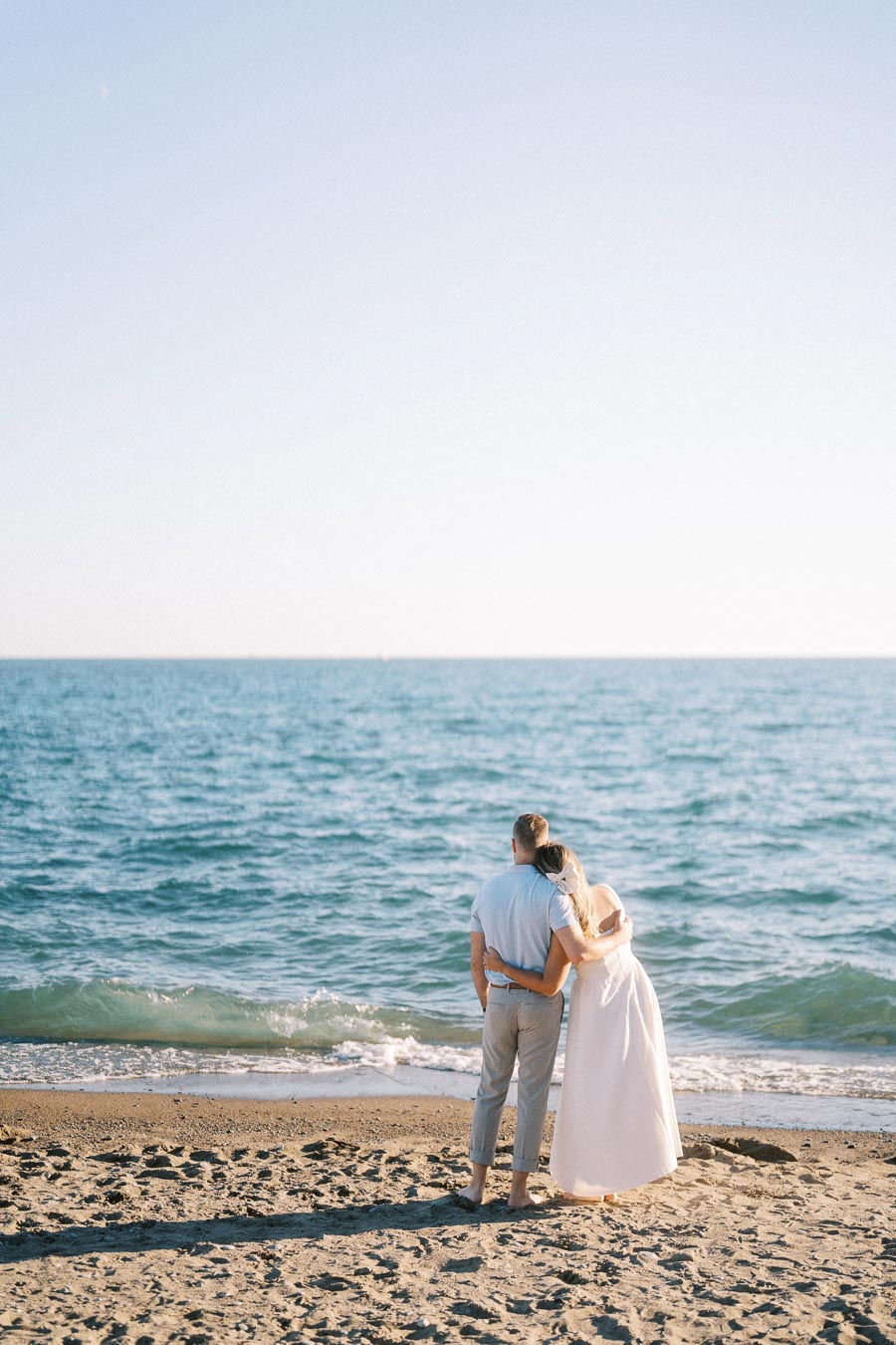 Couple embracing on a sandy beach, gazing at the calm ocean under a clear blue sky, symbolizing romance and tranquility.