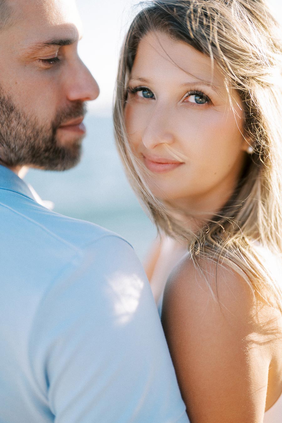 A couple embracing near the ocean, with the woman looking at the camera and the man gazing at her, capturing a romantic and serene beach moment.