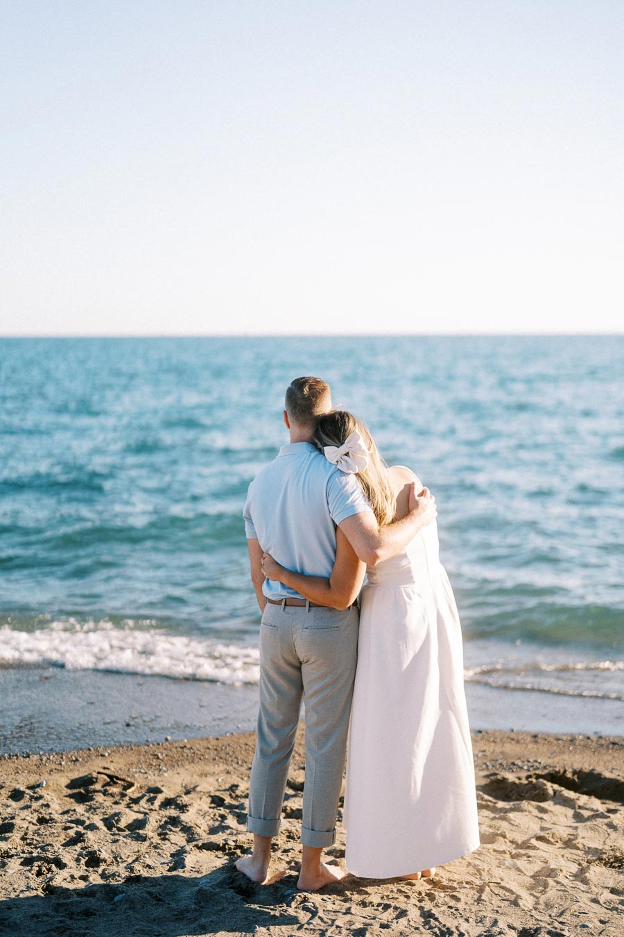 A couple embraces while standing barefoot on a sandy beach, gazing at the ocean under a clear blue sky, conveying a sense of romance and tranquility.