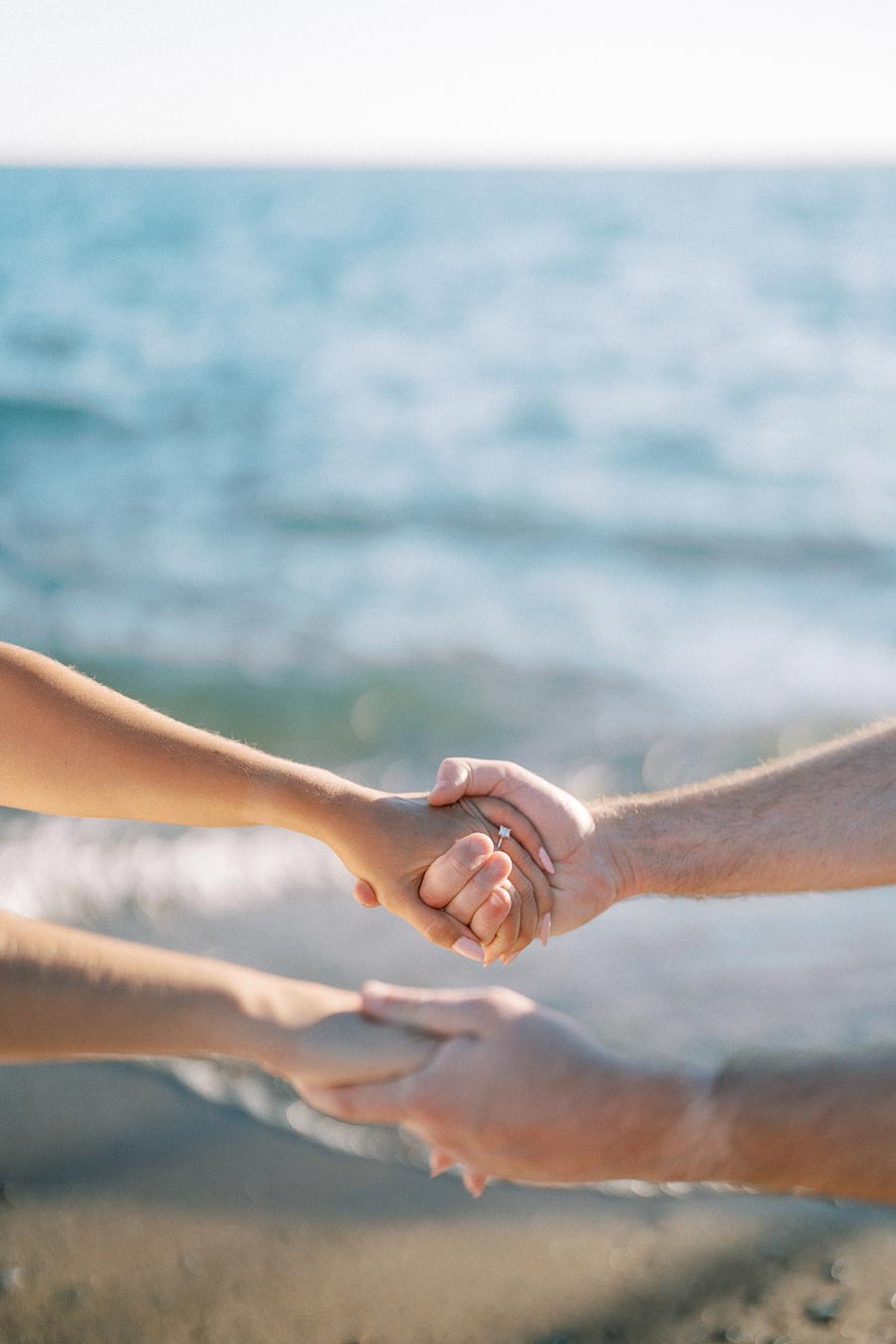 Close-up of a couple holding hands on a beach, with a sparkling diamond engagement ring visible, symbolizing love and commitment by the sea.