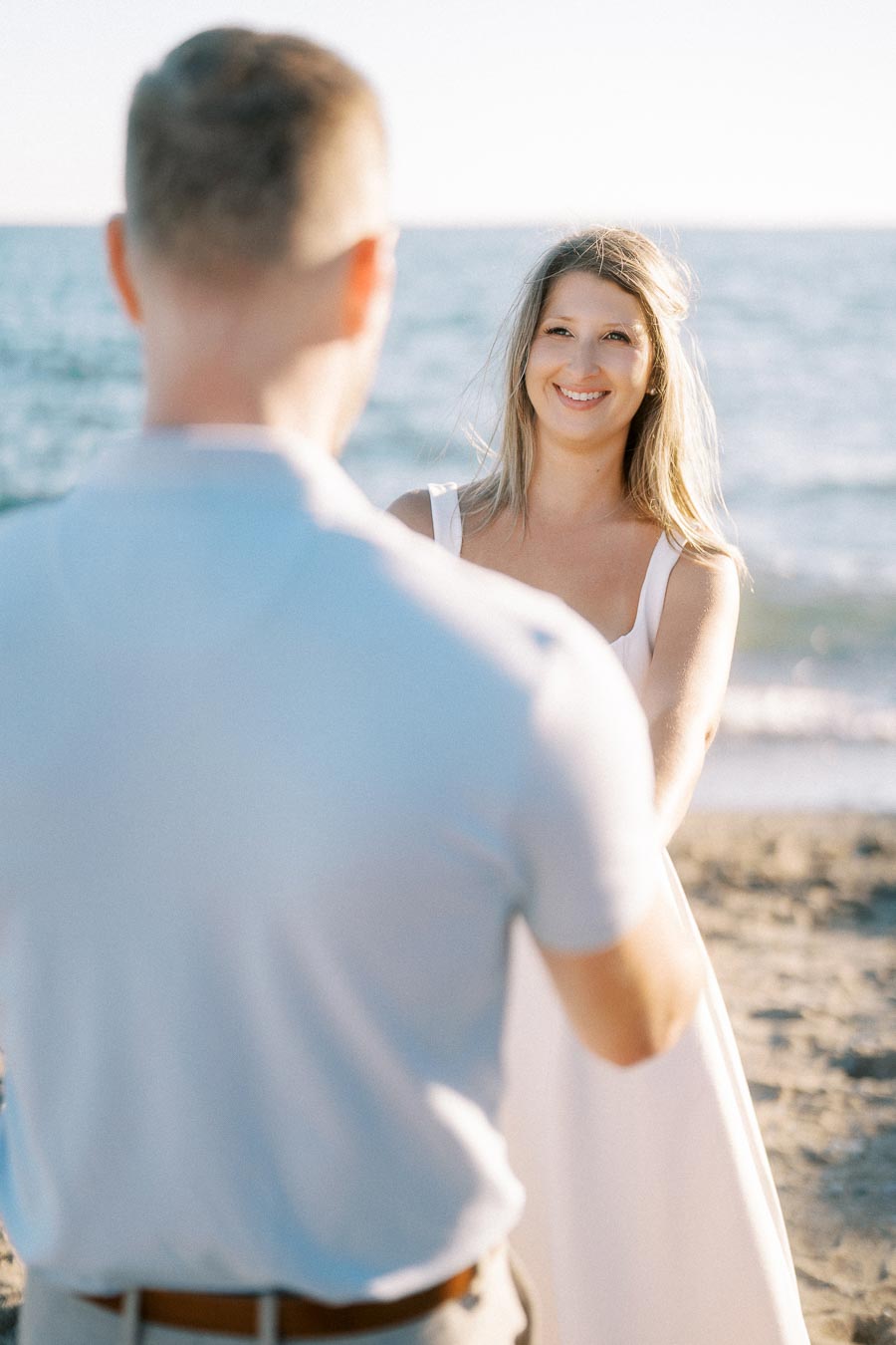 A couple holding hands on a sunny beach, with the ocean in the background; the woman is smiling warmly.