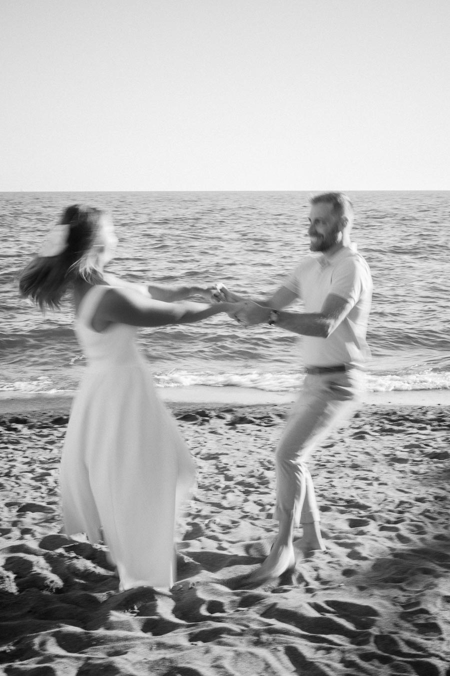 Black and white photo of a couple joyfully spinning on a sandy beach by the ocean, showcasing a romantic and energetic moment amidst the serene seaside backdrop.