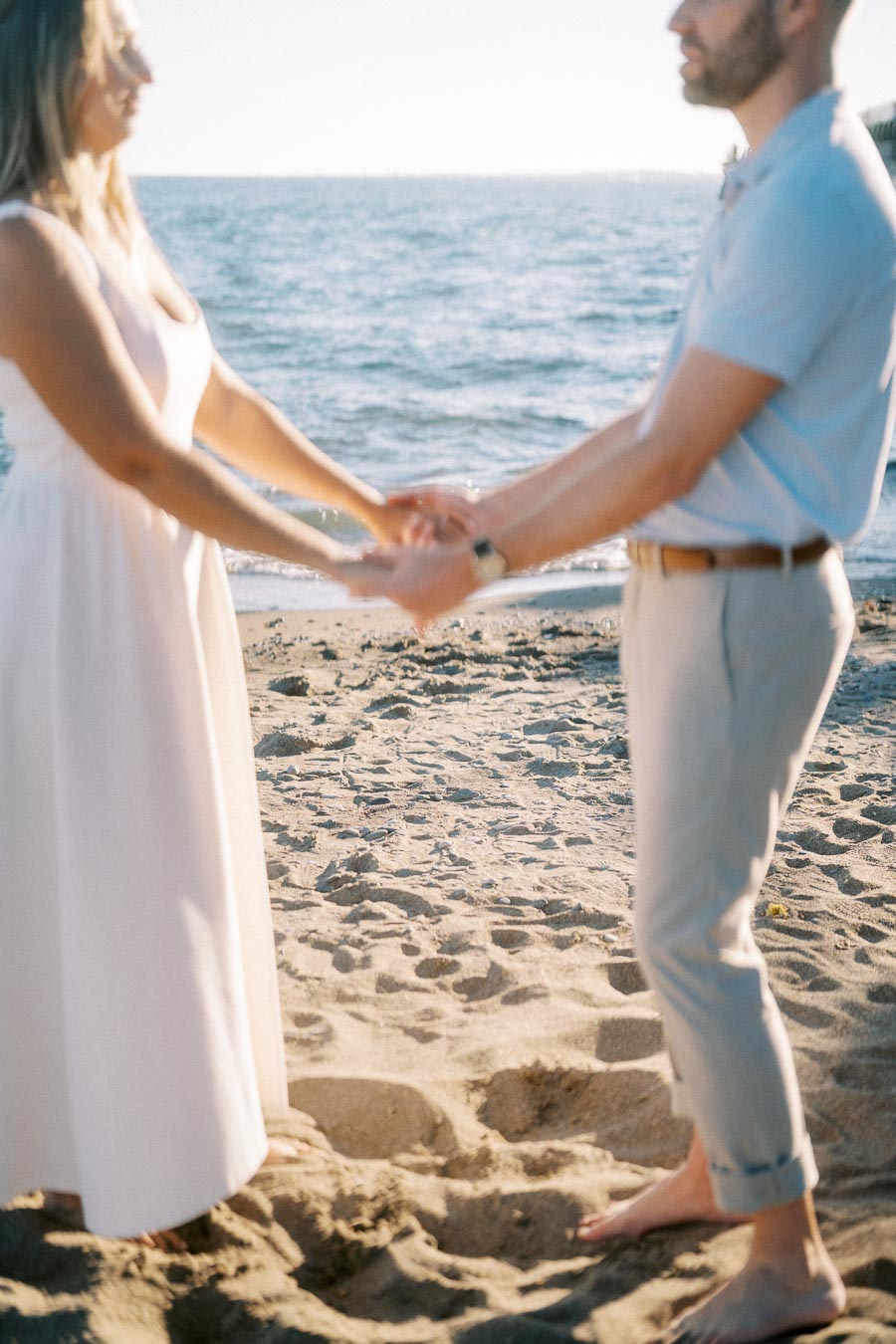 A couple holding hands on a sandy beach with the ocean in the background, wearing light-colored summer clothing.