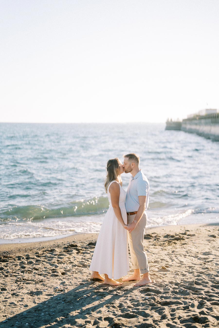 A couple kissing on a sunny beach with gentle waves, dressed in light summer clothes, holding hands and barefoot on the sand, with a pier in the background.