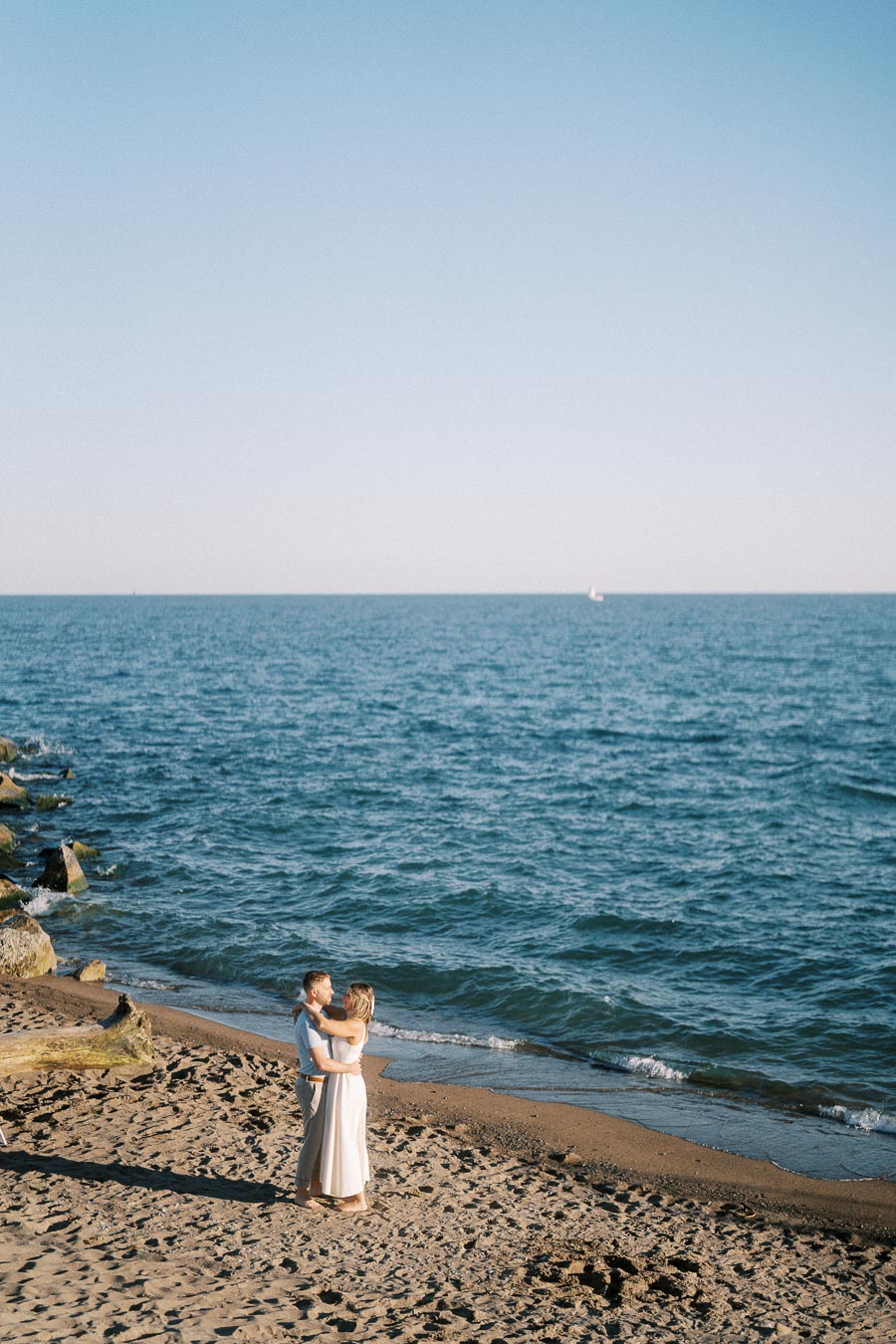 Couple embracing on a sandy beach with clear blue ocean in the background under a sunny sky, capturing a romantic seaside moment.