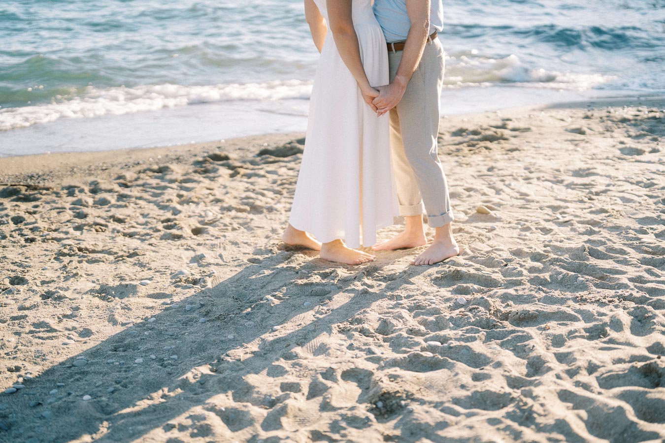 Couple holding hands barefoot on sandy beach near ocean waves, romantic seaside moment.