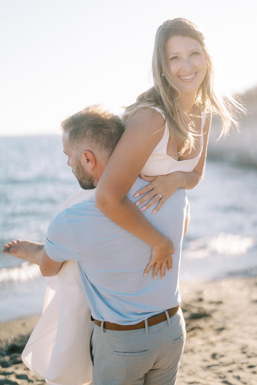 A couple enjoying a playful moment by the beach, with the woman in a white dress smiling while being carried by the man in a light blue shirt, capturing a romantic seaside memory.