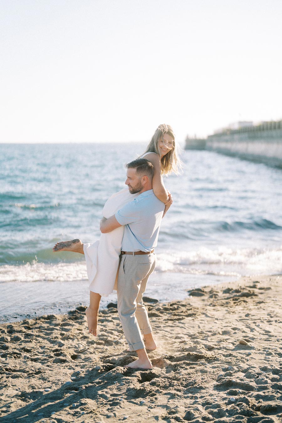 A couple enjoying a playful moment on a sandy beach at sunset, with the ocean in the background.