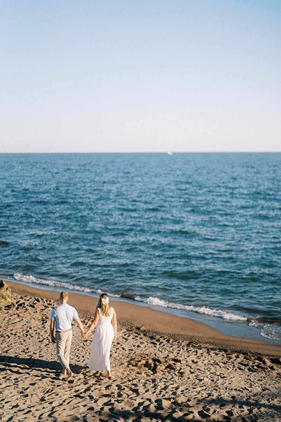 Couple walking hand in hand on a sandy beach towards the ocean under a clear blue sky, symbolizing romance and tranquility.