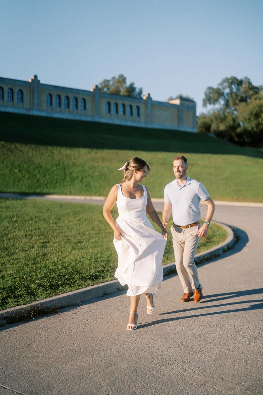 A happy couple enjoying a sunny day, walking along a paved path with green grass and a historic building in the background.