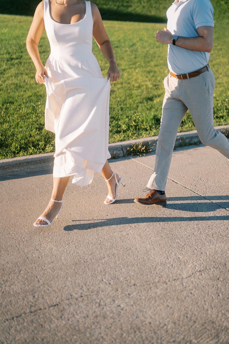 A couple joyfully running on a sunlit path; the woman is in a flowing white dress, and the man is dressed in light trousers and a blue shirt, with lush green grass in the background.