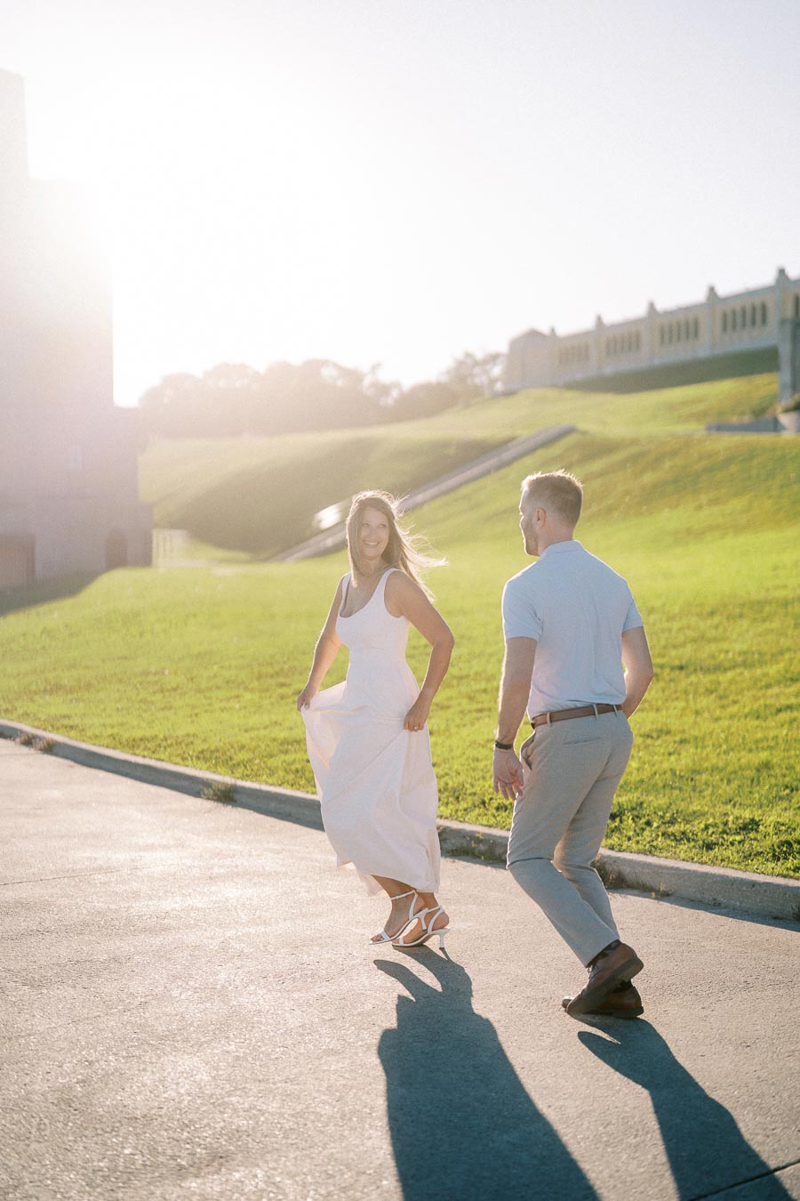 A couple joyfully walks along a sunlit path in a park, surrounded by lush green grass and historic architecture in the background.