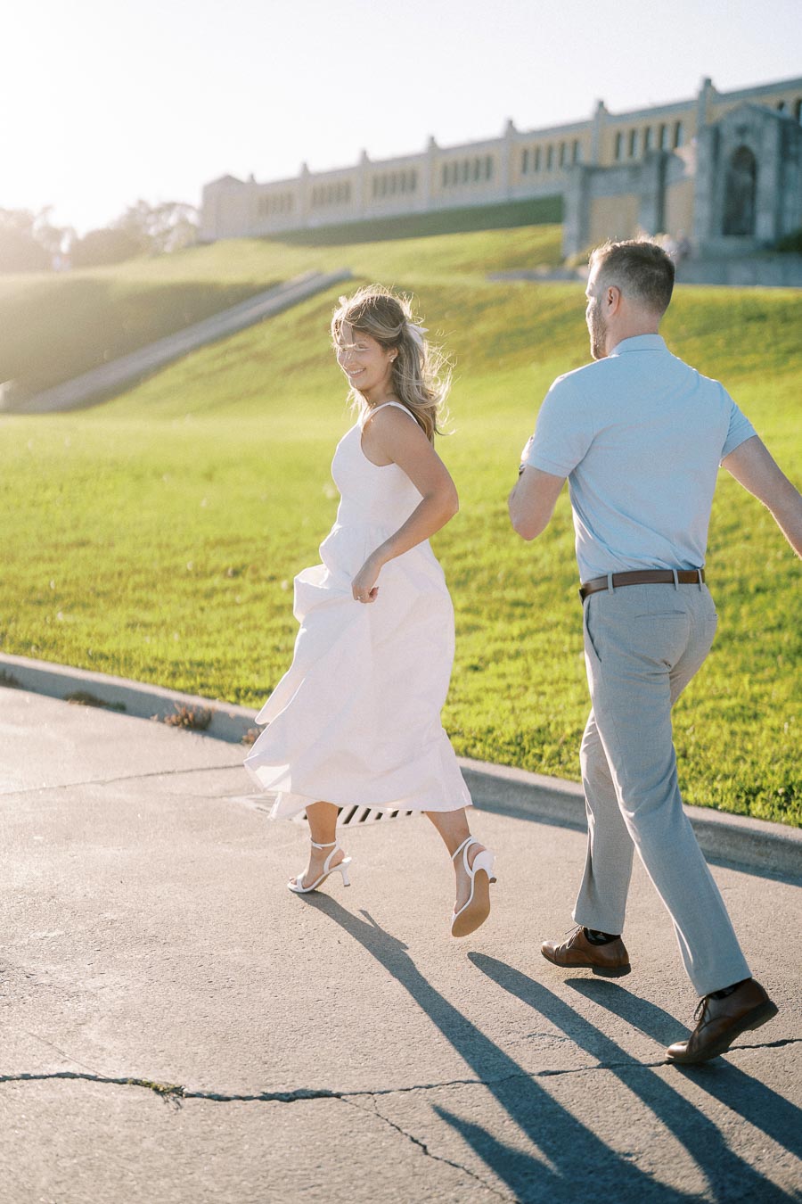 A couple joyfully running on a sunlit path with green grass and a historic building in the background. The woman wears a flowing white dress and the man is dressed in light colors, reflecting a bright, romantic atmosphere.
