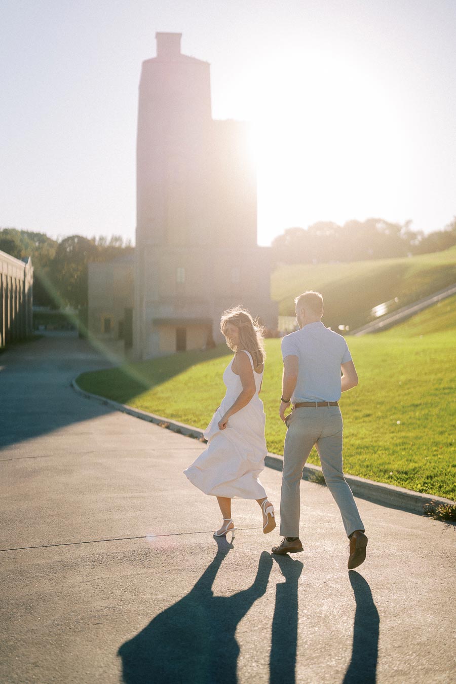 A couple joyfully running towards the sun on an outdoor path near a large tower and grassy area, with the sunlight creating long shadows.