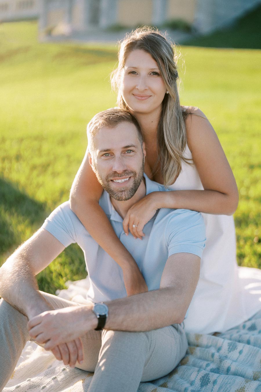 A couple sitting on a picnic blanket in a sunlit park, with the woman embracing the man from behind.