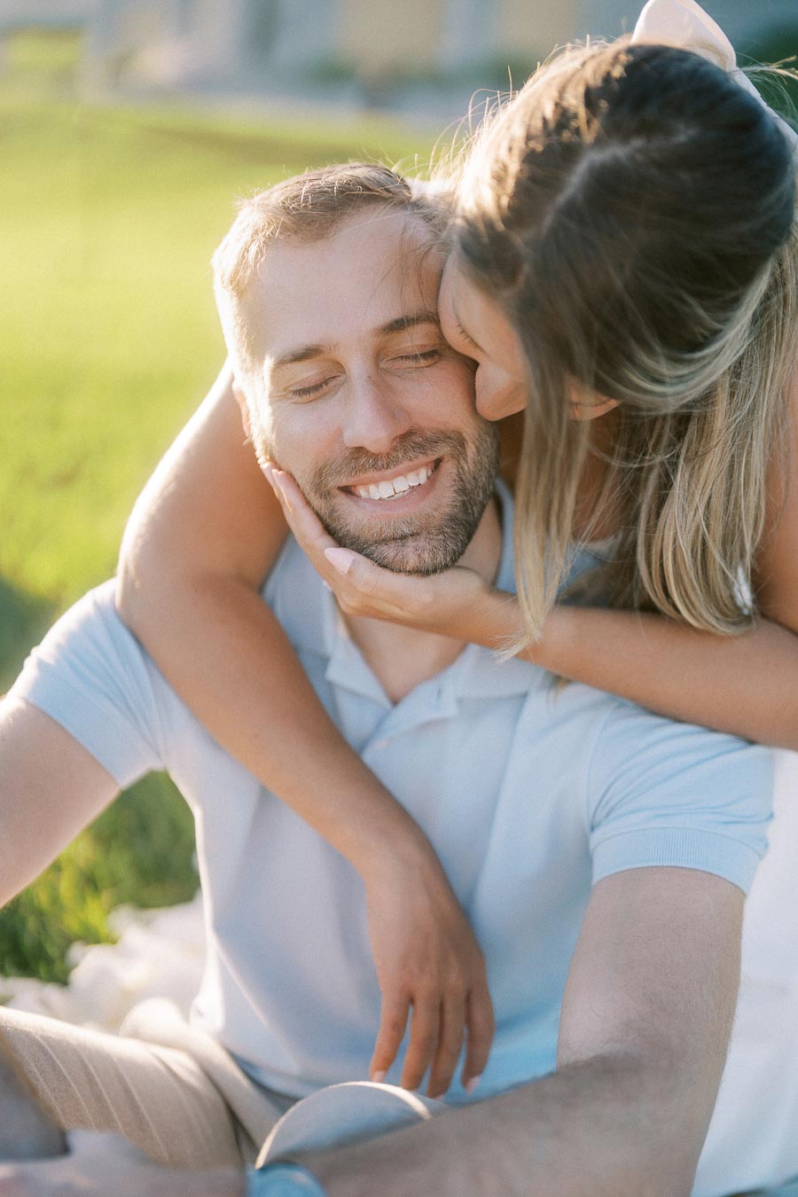 A happy couple embracing outdoors as the woman kisses the man's cheek, both smiling warmly under the sunlight, enjoying a romantic moment in a grassy park.