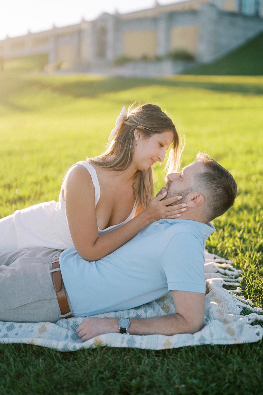 A couple enjoying a romantic picnic on a sunny day in a lush green park, with woman lovingly holding her partner's face while lying on a cozy blanket.