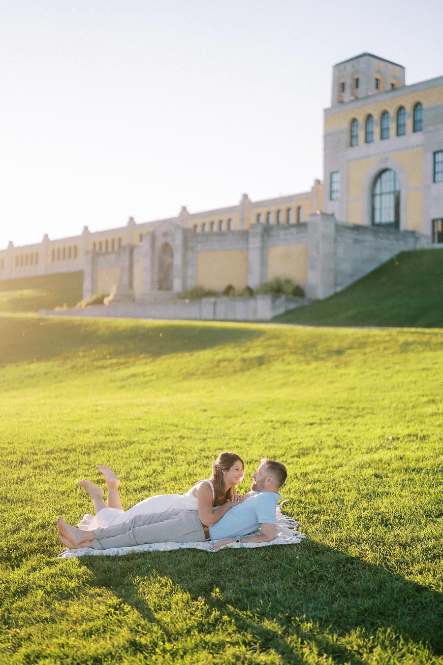 A couple lying on a blanket in a sunlit grassy field, with a historic building in the background, capturing a romantic outdoor moment during golden hour.