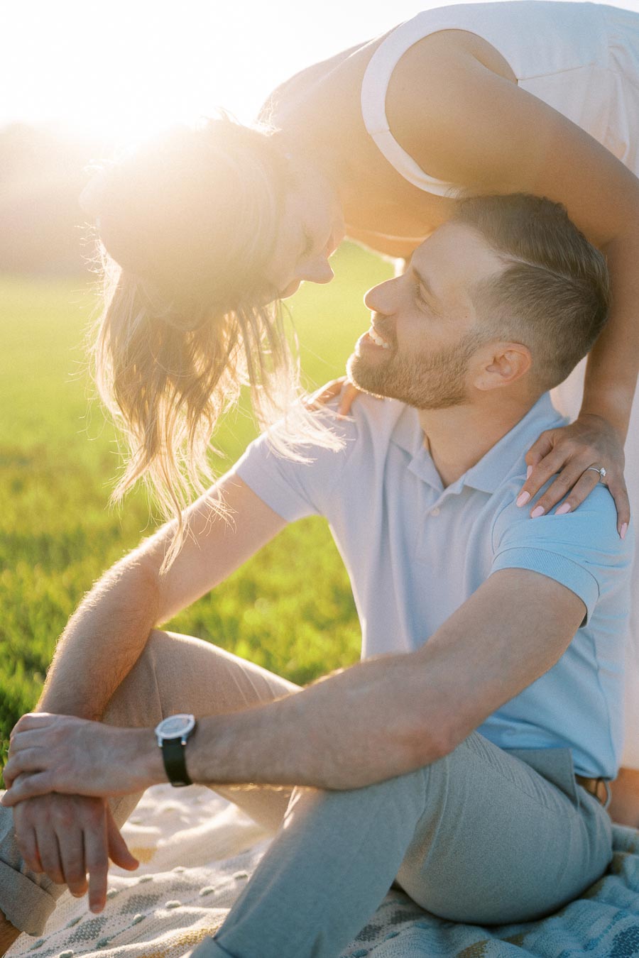 A couple enjoying a romantic moment in a sunny field, with the woman leaning over the man who is sitting on a picnic blanket, both smiling affectionately at each other.
