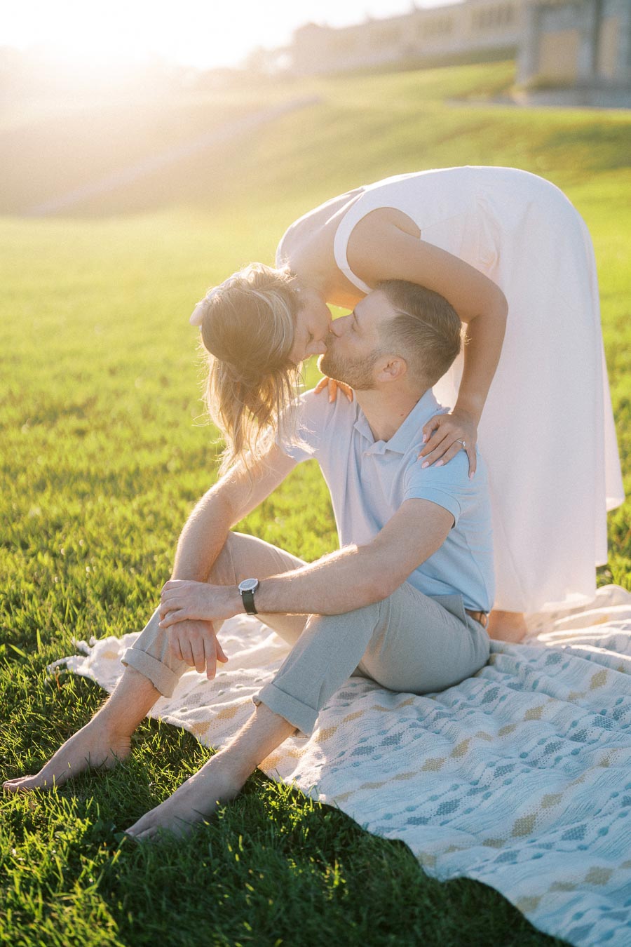 Romantic couple enjoying a tender moment during a sunny picnic in a green field, woman leaning down to kiss seated man on a blanket.