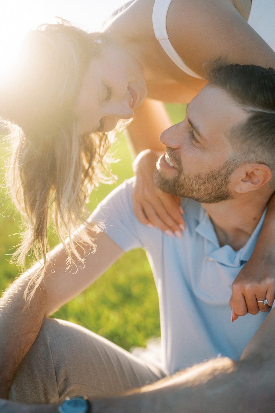 A couple smiling affectionately at each other in a sunlit park, with a woman embracing the man from behind while wearing a ring, symbolizing love and engagement.