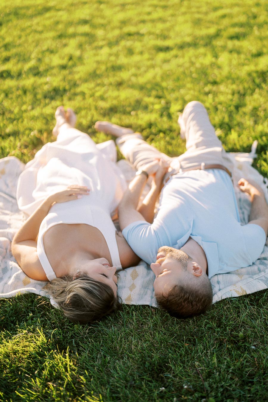 A couple lying on a blanket in a grassy field, holding hands and gazing lovingly at each other in a serene outdoor setting.