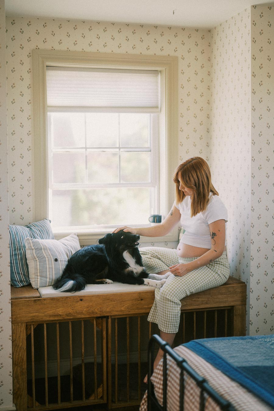 A woman with red hair and tattoos, seated in a cozy bedroom, gently petting a black and white dog on a window bench with striped cushions.