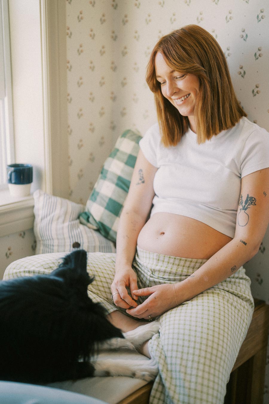 Pregnant woman with tattoos sitting on a window seat, smiling at a black dog, wearing comfortable attire in a cozy room.