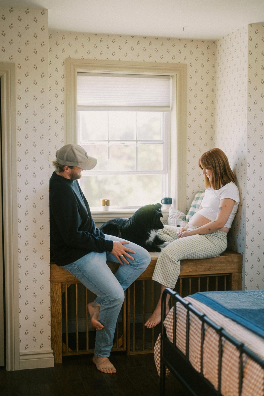 A couple sits in a cozy bedroom near a window, alongside a black and white dog. The scene includes soft natural light, patterned wallpaper, and a wooden bench, creating a warm, inviting atmosphere.