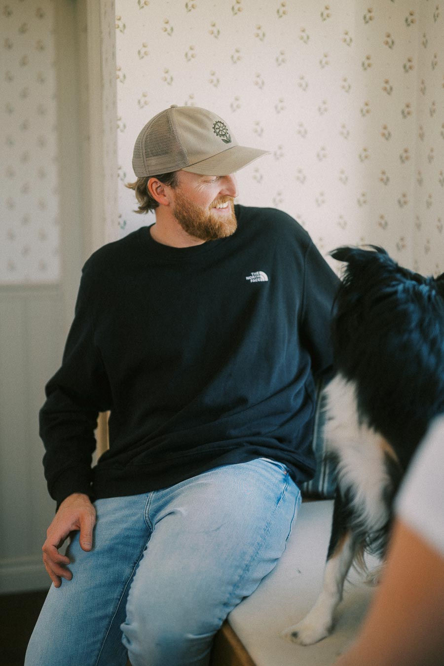 Man wearing a black North Face sweatshirt and beige cap smiling at a black and white dog indoors.