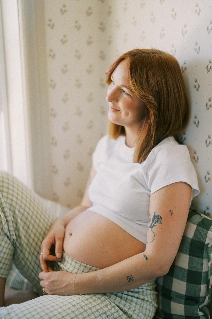 Pregnant woman in casual attire sitting by a window, gently holding her belly, looking content and relaxed.