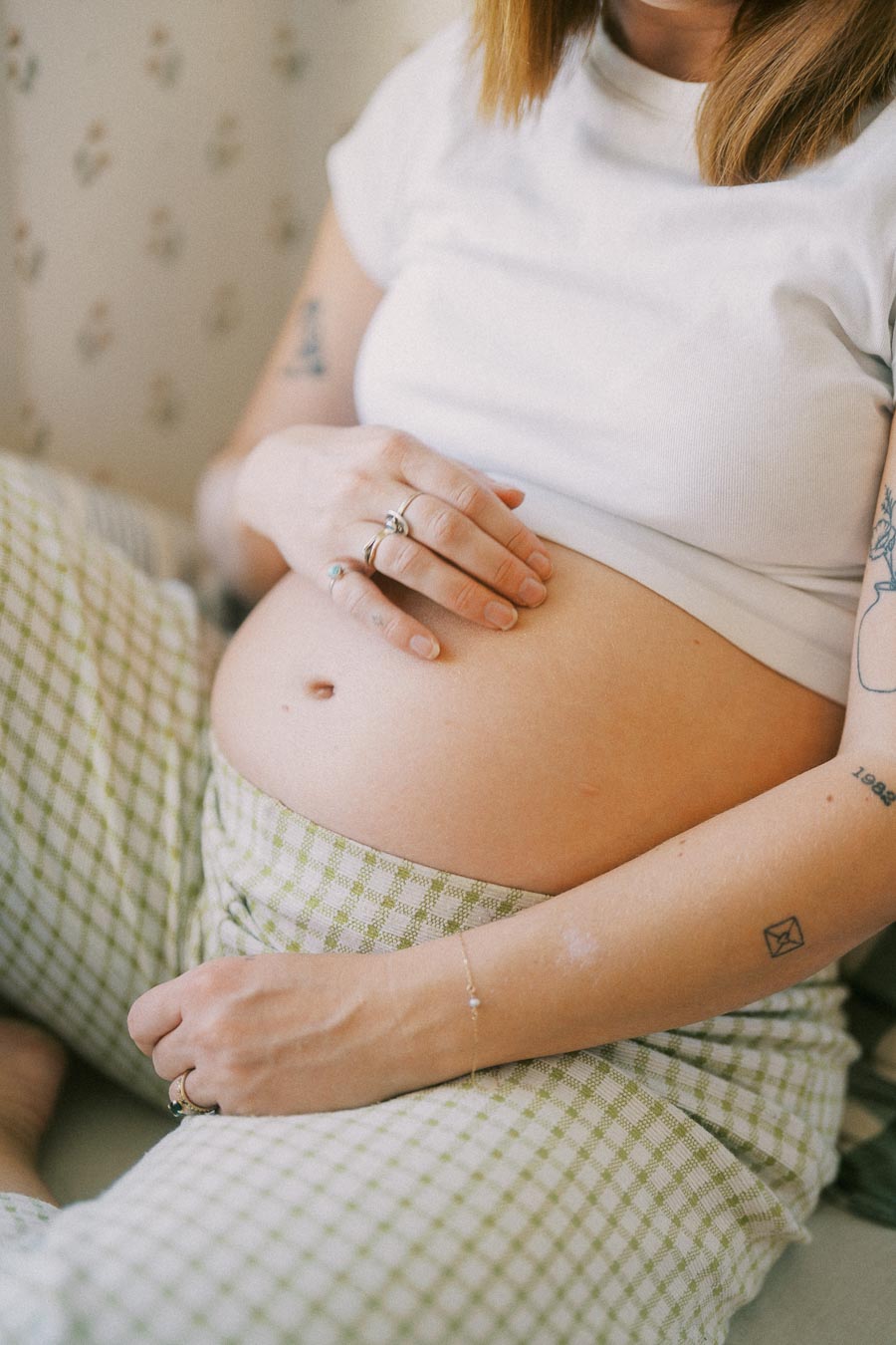 Pregnant woman gently cradling her belly, wearing a white t-shirt and checkered pants, with visible tattoos and jewelry, emphasizing maternal care and anticipation.