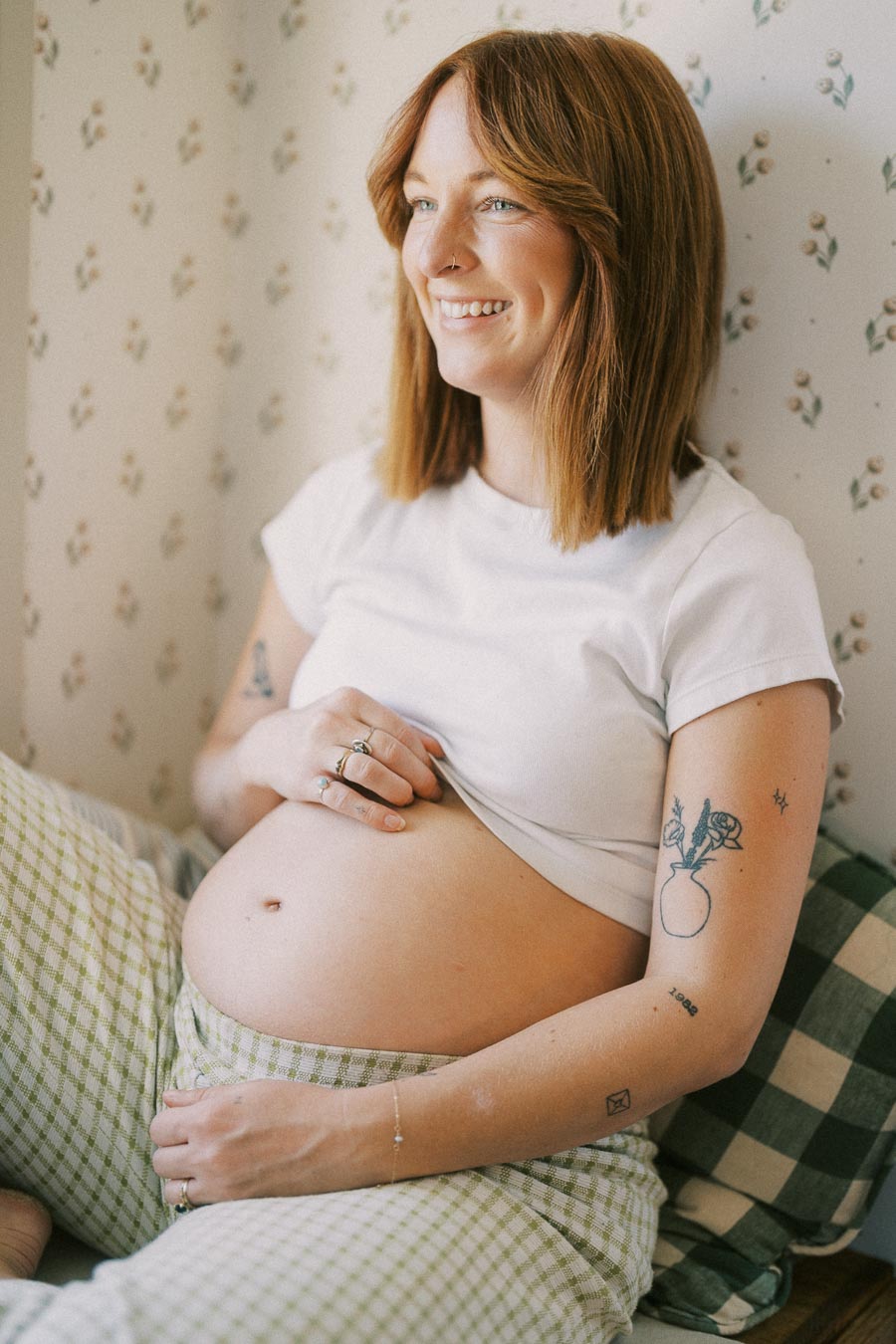 Smiling pregnant woman sitting by a wall with floral wallpaper, holding her belly and wearing a white t-shirt and checkered pants.